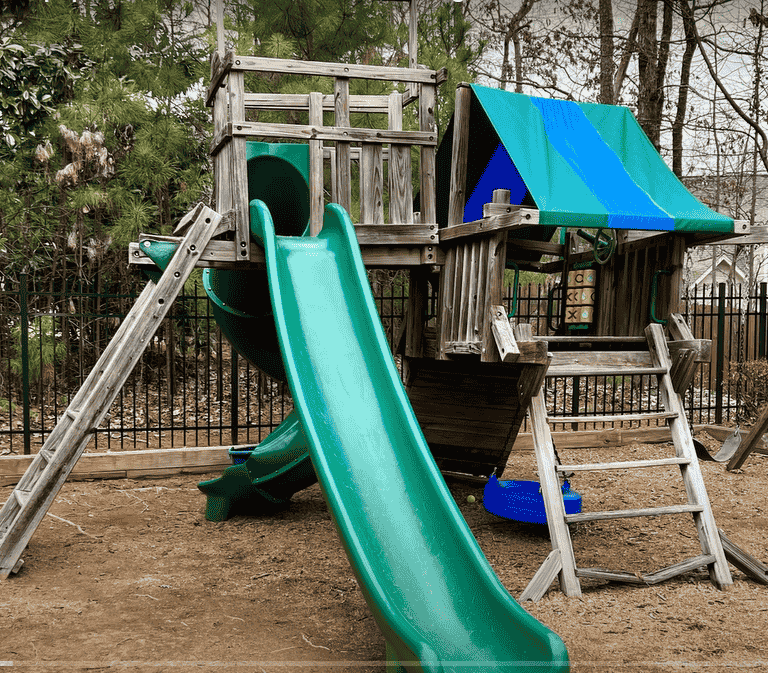 Playground items Demoltion : A wooden playground structure with a green slide, a blue tarp roof, and various climbing elements, set in a backyard with trees and a metal fence.