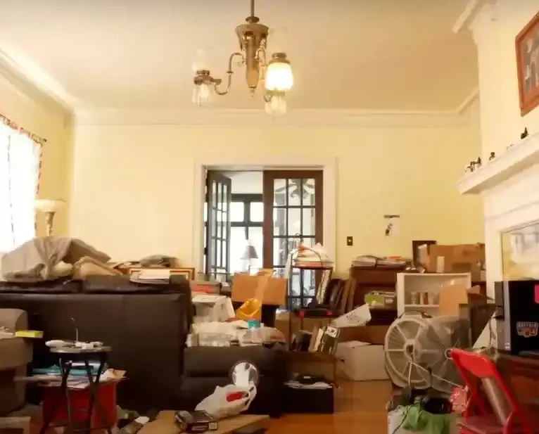 A cluttered living room with furniture, boxes, and various items scattered throughout. The room has high ceilings, a chandelier, and a fireplace.