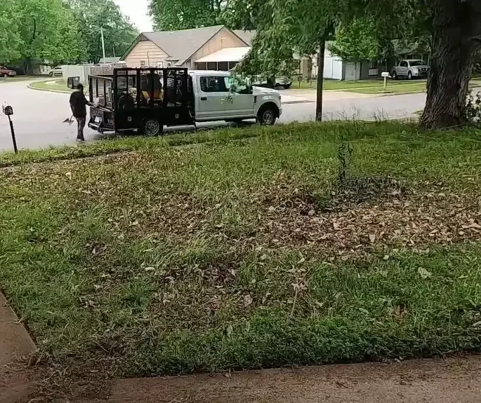 yard debris removal Wichita KS : A white pickup truck with a trailer is parked in a residential driveway. A man is walking towards the truck, carrying a rake. The lawn is covered in leaves.