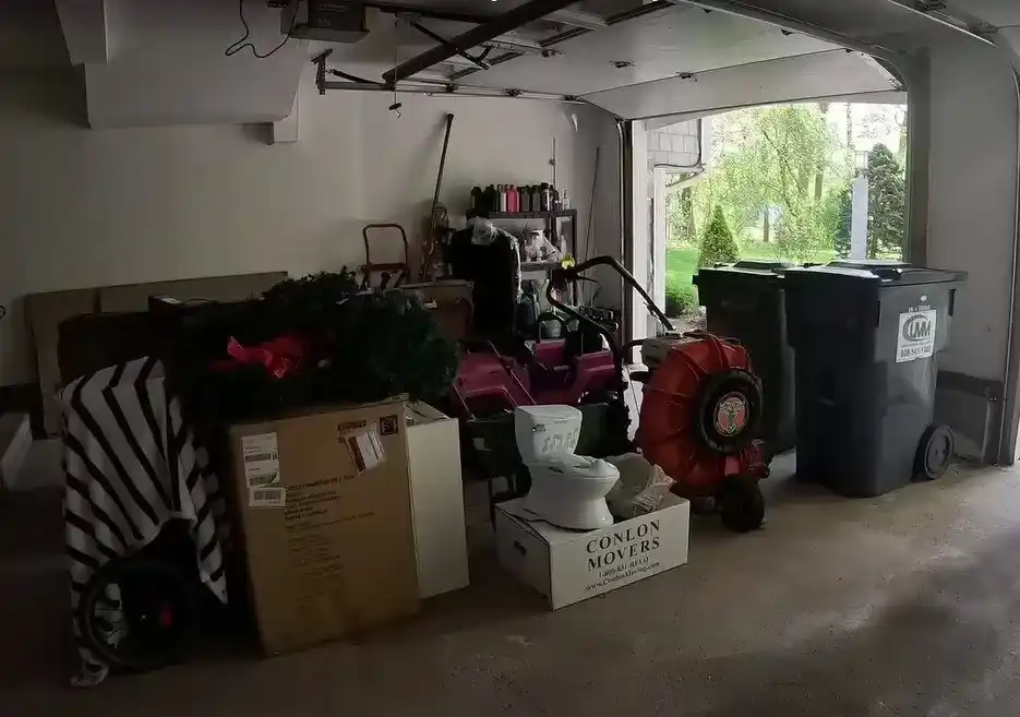 Garage Cleanout In Wichits KS : A cluttered garage filled with boxes, tools, and other miscellaneous items. There is a partially open garage door, a lawnmower, a leaf blower, and a trash bin visible in the background.