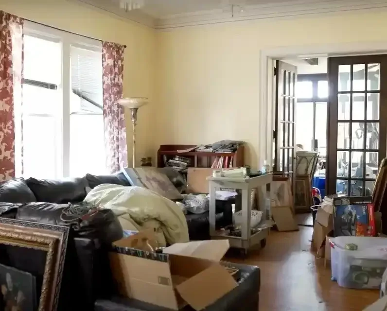 Estate Cleanout in Wichita KS: A cluttered living room with furniture, boxes, and various items scattered throughout. The room has high ceilings, a chandelier, and a fireplace.